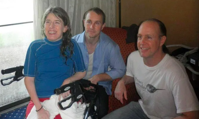 Family of three young people. A daughter in a blue shirt sitting on a walker. Two brothers crouching near her